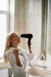 © JustLife - Young happy smiling woman wearing bathrobe using hair dryer in bathroom.