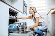 Beautiful caucasian blond housewife in apron crouching in front of dishwasher and putting dishes. Kitchen interior.
