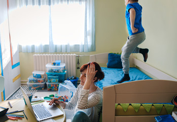 Wall Mural - Mother working remotely on laptop while taking care of her son playing with toys in his room.