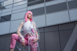 Young woman in pink pant suit and raincoat with pink hair on metal building wall background. Conceptual fashion photography. Copy space