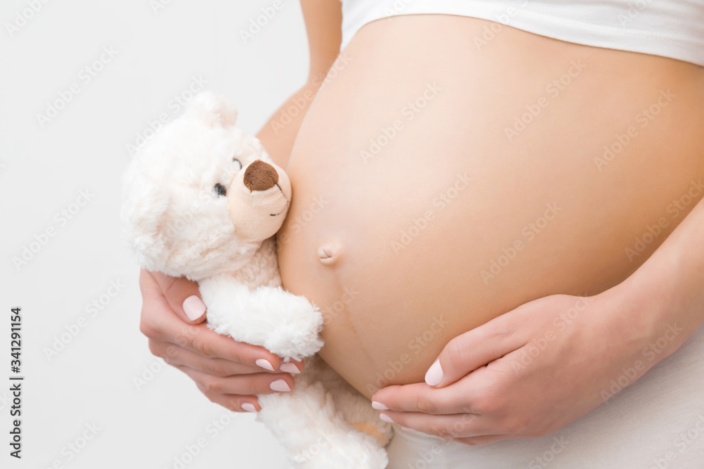 Hand holding smiling white teddy bear. Young woman naked belly. Emotional loving moment in pregnancy time - 30 weeks. Baby expectation. Love, happiness and safety concept. Closeup. Side view. - obrazy, fototapety, plakaty 