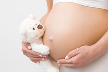 Hand Holding Smiling White Teddy Bear. Young Woman Naked Belly. Emotional Loving Moment In Pregnancy Time - 30 Weeks. Baby Expectation. Love, Happiness And Safety Concept. Closeup. Side View.