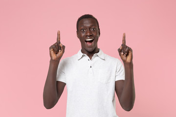 Surprised young african american man guy in white polo shirt posing isolated on pastel pink background studio portrait. People emotion lifestyle concept. Mock up copy space. Pointing index fingers up.