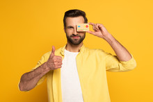 Smiling Man Covering Eye With Model Of Credit Card And Showing Thumb Up Gesture On Yellow Background