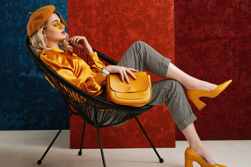 Full-length studio fashion portrait of elegant woman wearing yellow color sunglasses, beret, silk blouse, houndstooth printed trousers, pointed toe shoes, posing with stylish leather bag
