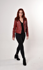 Portrait of a pretty girl with red hair wearing black jeans and boots with leather jacket.  full length standing pose on a studio background.