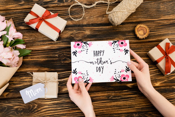 Wall Mural - cropped view of woman holding greeting card with happy mothers day lettering near presents and flowers on wooden surface