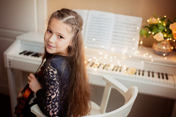 Wall Mural - Cute girl with a violin in her hands sits near the piano, bokeh from a garland. Music concept