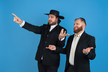Pointing, Inviting. Portrait Of A Young Orthodox Jewish Men Isolated On Blue Studio Background. Purim, Business, Festival, Holiday, Celebration Pesach Or Passover, Judaism, Religion Concept.