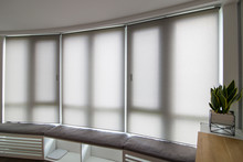 Motorized Roller Shades In The Interior. Automatic Roller Blinds Beige Color On Big Glass Windows. Home Luxury Curtaines Are Above The Windosill With Pillows. Summer. Green Trees Outside.