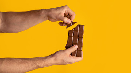 Wall Mural - Closeup of man breaking piece of chocolate from bar, orange background. Copy space. Panorama