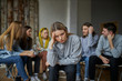 © Roman - young female suffer from alcohol addiction, sit apart from group of people gathered in the club of anonymous alcoholics
