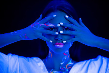 young caucasian woman with fluorescent body art closing eyes with hands, look at camera through fingers. blue neon light on her face. isolated dark background