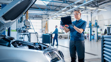 Female Mechanic Uses A Tablet Computer With An Augmented Reality Diagnostics Software. Specialist Inspecting The Car In Order To Find Broken Components Inside The Engine Bay. Modern Car Service.