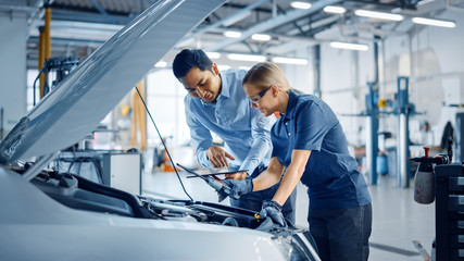 Wall Mural - Instructor with a Tablet Computer is Giving a Task for a Future Mechanic. Female Student Inspects the Car Engine. Assistant is Checking the Cause of a Breakdown in the Vehicle in a Car Service. 