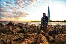 Man Sitting On Rock By Sea Against Sky During Sunset