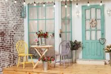 Garden Table And Chairs On The Summer Porch Of The House. Summer Terrace With Retro Light Bulbs Garlands. Beautiful Porch With Spring Decorations In The Photo Studio