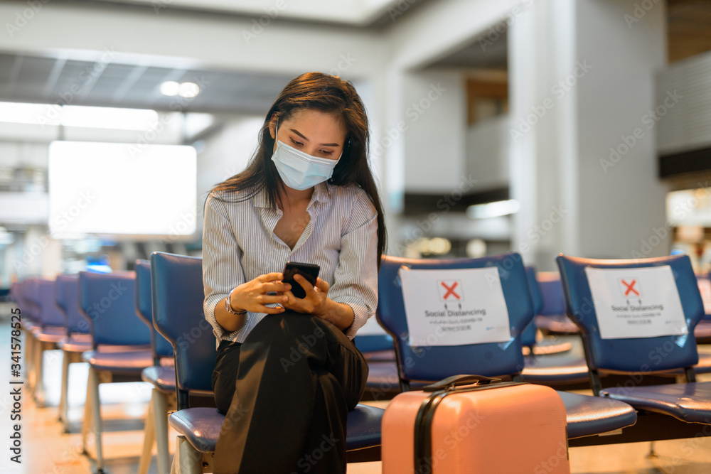 Young Asian tourist woman with mask using phone and sitting with distance at the airport - obrazy, fototapety, plakaty 