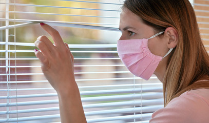 Wall Mural - Young woman in pink home made cotton virus face mask, looking through window blinds outside. Quarantine or stay at home during coronavirus covid-19 outbreak