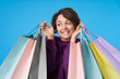 © timtimphoto - Positive young pretty dark haired curly woman in purple sweatshirt keeping shopping bags in raised hands and looking cheerfully aside, standing over blue background