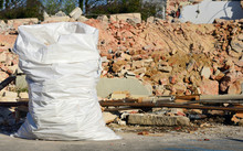 A Large White Plastic Sack Full Of Rubble Stands In Front Of A Demolished House With Red Bricks In The Background