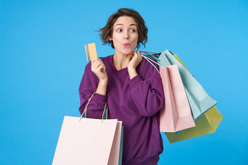 Wall Mural - Portrait of young positive short haired curly woman folding cheerfully her lips while looking aside and keeping shopping bags and credit card in raised hands, isolated over blue background