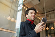 © timtimphoto - Pleasant looking young attractive dark haired man smiling gladly while keeping smartphone in raised hand, posing over cafe interior in elegant clothes