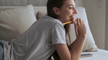 Poster - Side view of Pensive happy pretty brunette woman holding pencil and looking away while lying on sofa at home