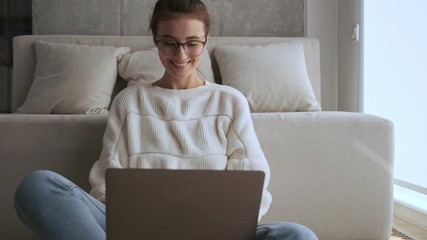 Poster - Cheerful pretty brunette woman in eyeglasses using laptop computer and looking at the camera after that while sitting near the window at home