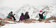 © guruXOX - Group of female and male snowboarder and skier, sit and rest on the rock in the snow, and enjoy in beautiful landscape of panoramic Alps mountain range viewpoint. Back view. French Alps, Les 2 Alpes