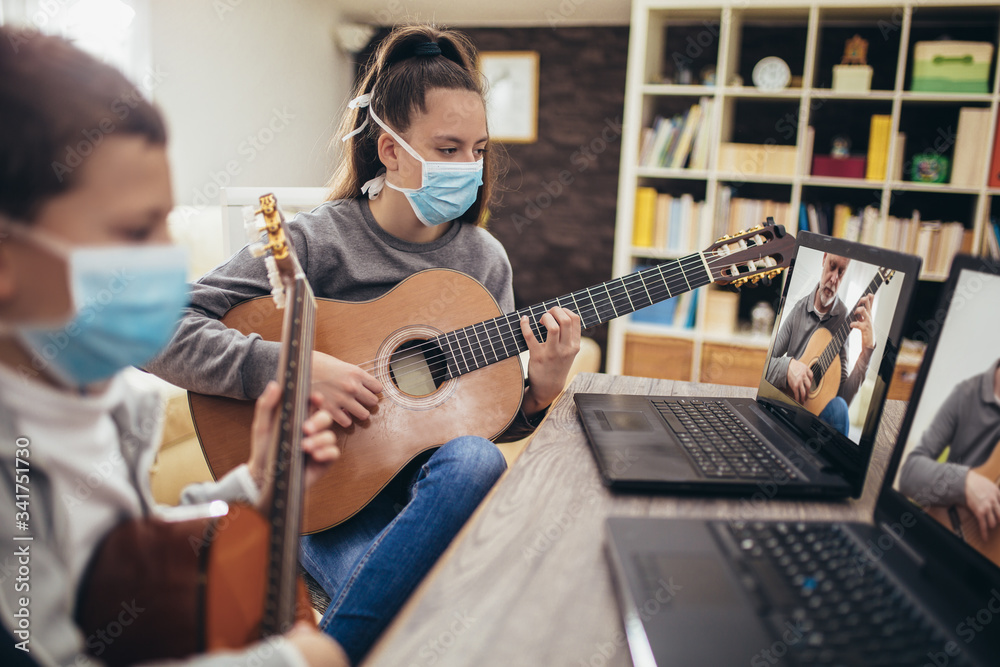 Boy and girl, wear protective masks, playing acoustic guitar and watching online course on laptop while practicing at home. Online training, online classes. - obrazy, fototapety, plakaty 