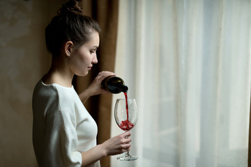 Young woman pours red wine into a large glass near a curtained window