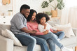 © Prostock-studio - Happy black parents and little daughter relaxing with smartphone at home