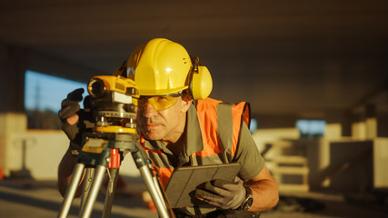 Inside of the Commercial / Industrial Building Construction Site: Professional Engineer Surveyor Takes Measures with Theodolite, Using Digital Tablet. In Background Skyscraper Formwork Frames Crane 