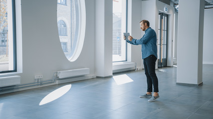 Wall Mural - Young Hipster Man in Glasses Standing in Empty Office and Map it with an Augmented Reality Software on a Tablet. Sunlight Shines Through Big Windows.