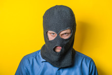 Man Close Up Thief In A Mask And A Blue Shirt On A Yellow Background Looks Slyly To The Camera. Mimicry. Gesture. Photo Shoot/ Evil Criminal Wearing Balaclava