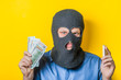 © aeroking - man close up thief in a mask and a blue shirt on a yellow background looking slyly at the camera, holding the money and the phone. Mimicry. Gesture. photo Shoot