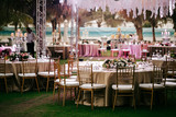 Fototapeta  - International Wedding outdoor celebration EVENING party under palm trees. Served tables on green area in hotel. Landyard. Beige and pink colors. Close-up and wide angle.