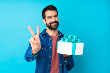 Young handsome man with a big cake over isolated blue background smiling and showing victory sign