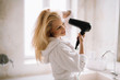 © JustLife - Young happy smiling woman wearing bathrobe using hair dryer in bathroom.