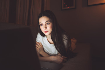 Woman is looking on a laptop screen, lying on a sofa at home in the dark. Watching video, chatting with friends 