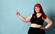 Overweight redhead woman in red headband, black top and leggings. She pointing at something, posing on blue studio background