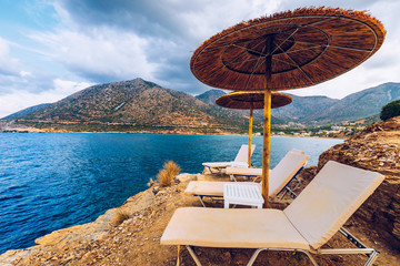 Wall Mural - Summer vacation destination. Straw sunshades and sunbeds on the empty rocky beach with sea in the background. Vacation And Tourism Concept. Sunbeds On The Paradise Beach. Umbrellas and sunbeds.