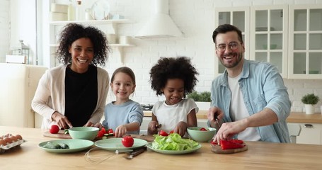 Wall Mural - Cheerful mixed race family with kids cooking together, laughing, looking at camera in kitchen. Cute african and caucasian children daughters helping happy parents preparing healthy vegetable salad.