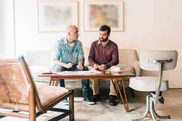 Interior designer and owner talking about sketches, drawings. Focused men with papers sitting in living room at home and talking. Renovation concept