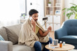 people, health and fever concept - sick indian man in blanket with thermometer calling on smartphone at home