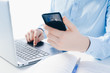 © Nataliya - a man in a blue shirt is working on a laptop, holding a smartphone in his hands. Remote work, remote control, online stores