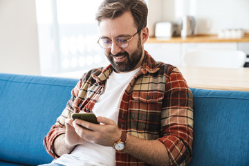 Sticker - Portrait of young man smiling and using cellphone on sofa at home