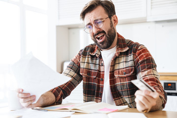 Portrait of shocked man holding credit card and papers while working