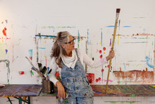 Proud Older Artist Woman, In Her Fifties With Grey Hair And Black Glasses Holds A Big Paintbrush